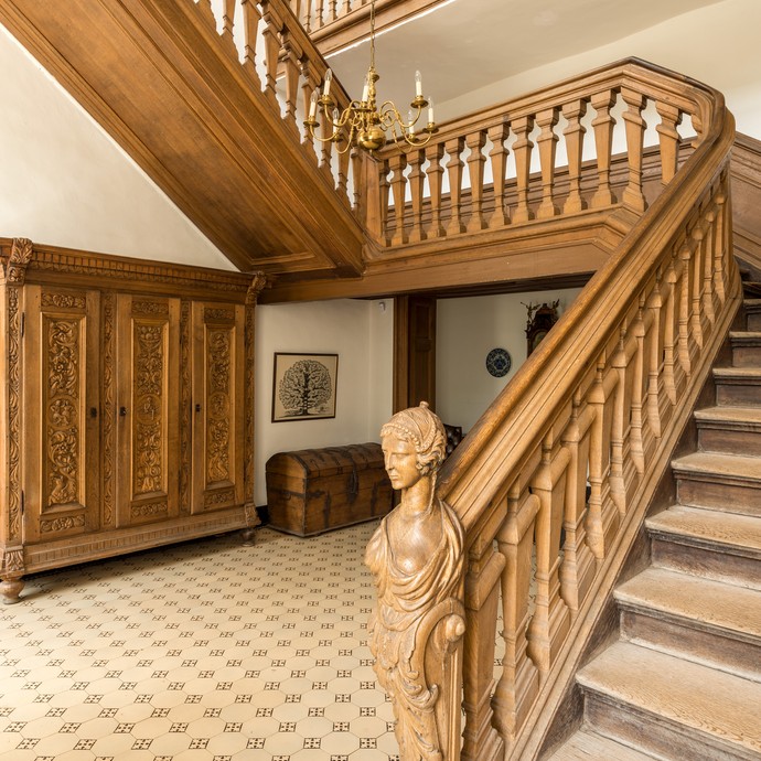 Große braune Holztreppe mit dem geschnitzten Kopf von Clara F. Antoinette Gräfin von Westerholt-Lembeck. Sie hat den Bau beauftragt. (vergrößerte Bildansicht wird geöffnet)