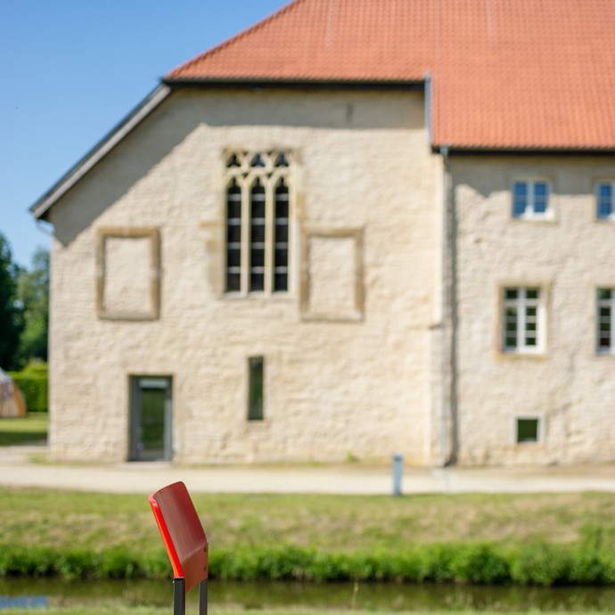 Ein roter Stuhl auf einer Wiese steht im Vordergrund. Dahinter ist verschwommen das Klostergebäude zu erkennen. (vergrößerte Bildansicht wird geöffnet)
