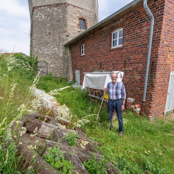 Josef Deitermann steht vor einem roten Steingebäude. Vor ihm liegt ein großes Getriebezahnrad der Mühle. (vergrößerte Bildansicht wird geöffnet)