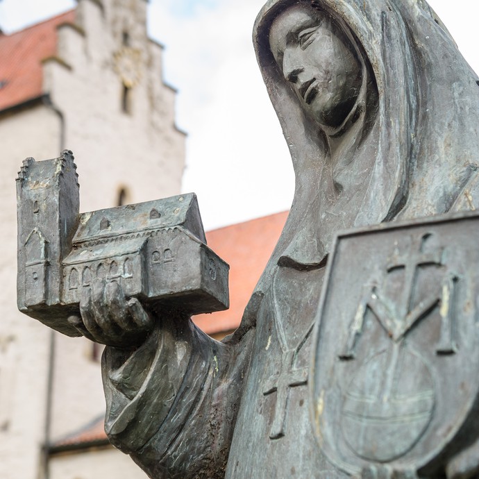Statue einer Frauenfigur mit einer Kirche in der Hand. Im Hintergrund steht die katholische Pfarrkirche St. Cornelius und Cyprianus in Metelen. (vergrößerte Bildansicht wird geöffnet)