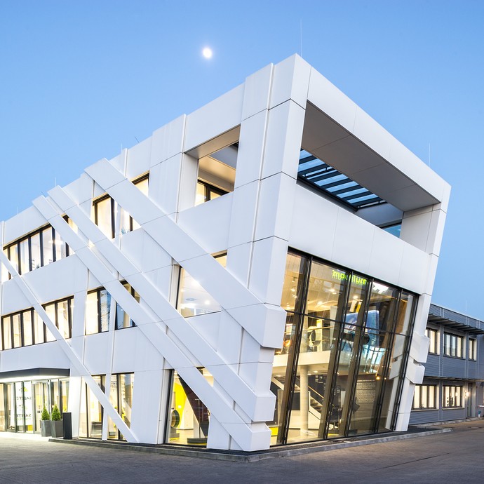 Außenansicht des bemerkenswerten Firmengebäudes in der Dämmerung. Die fensterreiche Fassade steht schräg. (vergrößerte Bildansicht wird geöffnet)