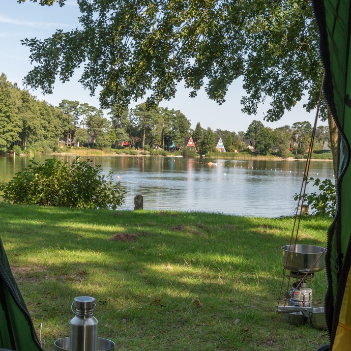 Das Foto zeigt den Blick aus einem Zelt auf den See. Am Rand ist die bunte Zeltplane, in der Mitte die grüne Rasenfläche und der See. (vergrößerte Bildansicht wird geöffnet)