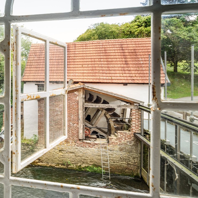 Blick durch ein Fenster auf ein Gebäude, dem ein Teil der Außenwand fehlt. Deshalb sieht man darin ein Mühlrad aus Holz. (vergrößerte Bildansicht wird geöffnet)