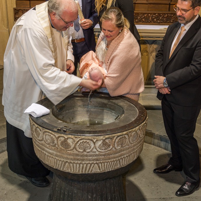 Am mit Mustern verzierten Taufstein wird ein Baby vom Pfarrer getauft. (vergrößerte Bildansicht wird geöffnet)