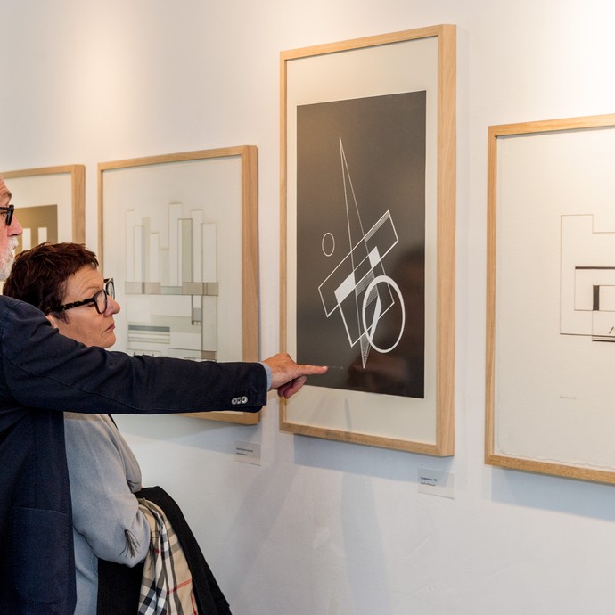 Ein Mann und eine Frau schauen sich Bilder der Ausstellung an. Der Mann zeigt auf ein schwarz-weißes Bild, das an der Wand hängt. (vergrößerte Bildansicht wird geöffnet)