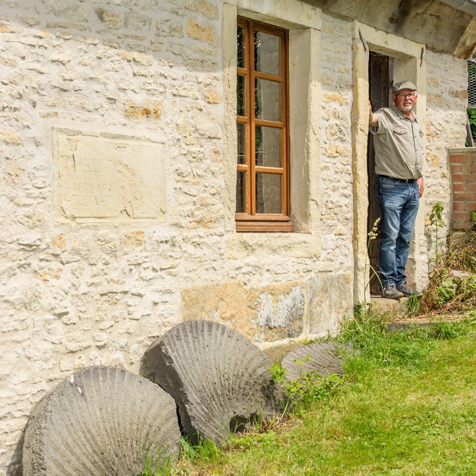Sandsteingebäude mit Rasenfläche und einem alten zerbrochenen Mahlstein. In der Tür des Hauses steht der Eigentümer. (vergrößerte Bildansicht wird geöffnet)