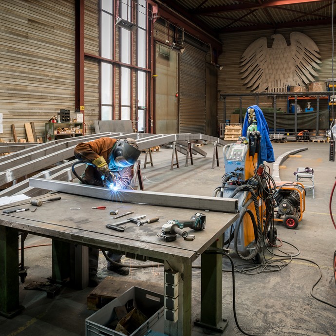 Eine große Halle mit langen Metallstücken. An einem Tisch mit Werkzeugen bearbeitet ein Arbeiter ein Metallstück. (vergrößerte Bildansicht wird geöffnet)