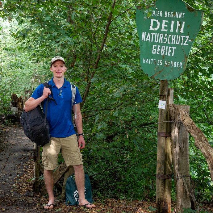 Ein junger Mann steht im Wald neben einem Schild mit der Aufschrift "Hier beginnt Dein Naturschutzgebiet. Halte es sauber". (vergrößerte Bildansicht wird geöffnet)