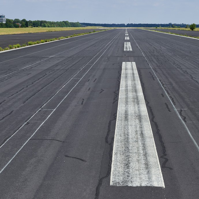Startbahn eines Flughafens mit dunkelgrauem Boden und weißen Wegstreifen. Im Hintergrund sind grüne Wiesen und Bäume. (vergrößerte Bildansicht wird geöffnet)