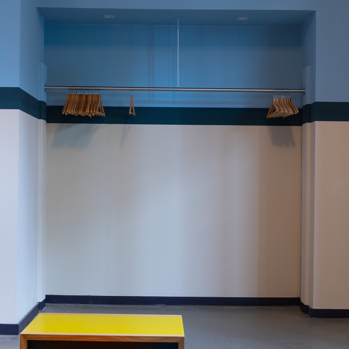 Leere Garderobe mit Kleiderbügeln aus Holz. Vor der blauen Wand sind drei Hocker mit bunten Sitzflächen und hölzernen Beinen. (vergrößerte Bildansicht wird geöffnet)