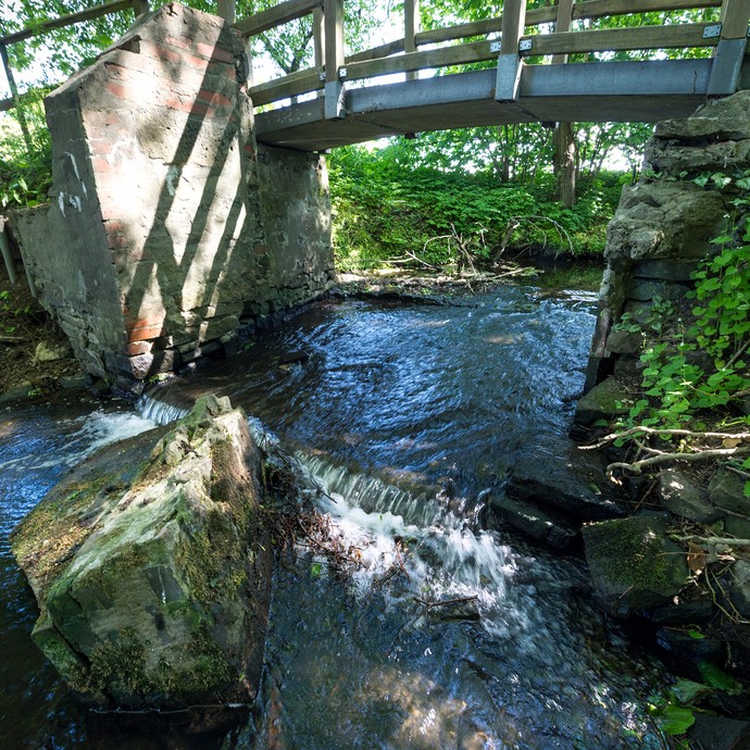 Eine Brücke führt über den Mühlenbach. Im Vordergrund ist eine kleine Stromschnelle. (vergrößerte Bildansicht wird geöffnet)