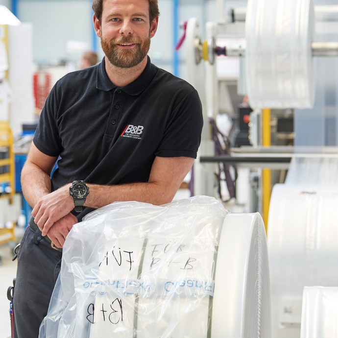 Der Mitarbeiter Christopher Stork-Bohmann lehnt sich an eine Folienrolle an der Verpackungsmaschine. Er blickt in die Kamera und lächelt. (vergrößerte Bildansicht wird geöffnet)