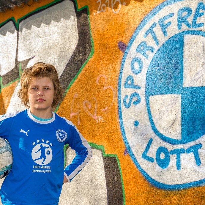 Der Jugendspieler Sami Rogatz steht im Trikot und mit Ball im Arm neben einer Graffitiwand mit dem blau-weißen Vereinslogo. (vergrößerte Bildansicht wird geöffnet)