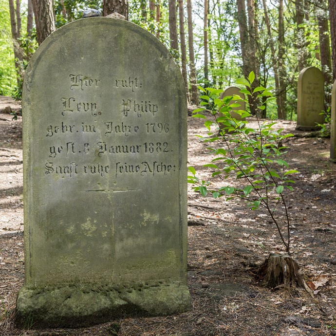 Ein sehr alter Grabstein auf dem jüdischen Friedhof. Die Schrift ist noch lesbar. Im Hintergrund stehen weitere Steine und Laubbäume. (vergrößerte Bildansicht wird geöffnet)