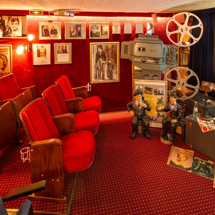Blick in den kleinen Kinosaal mit den typischen roten Kinosesseln. Rechts ist die Kinoleinwand. An der Wand hängen Filmplakate und Bilder. (vergrößerte Bildansicht wird geöffnet)