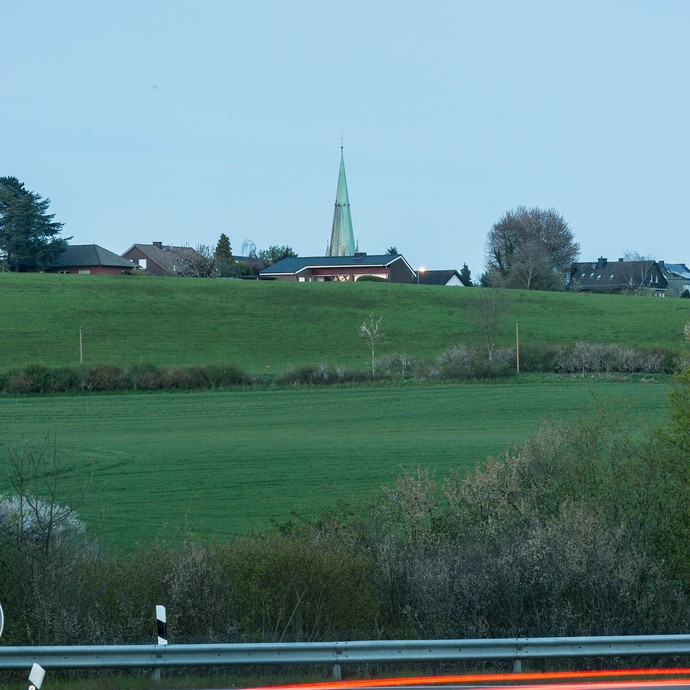 Hinter einer Anhöhe mit grüner Wiese erscheint der Kirchturm von St. Johannes Baptist. (vergrößerte Bildansicht wird geöffnet)