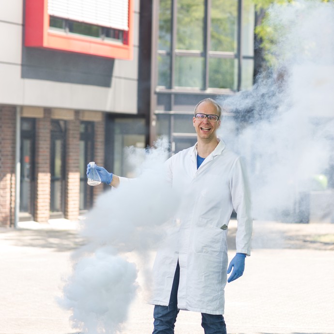 Dr. Thomas Muesmann ist Chemiker der Firma. Er steht vor der Firma. Neben ihm steigt Rauch aus einem Behälter auf. (vergrößerte Bildansicht wird geöffnet)