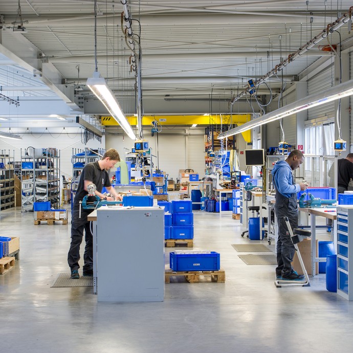 In einer großen Halle sind viele blaue Kisten mit Materialien. Zwei Arbeiter stehen an Werkbänken und bauen etwas zusammen. (vergrößerte Bildansicht wird geöffnet)