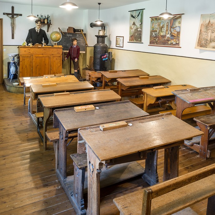 Viele Holzbänke stehen hintereinander. Vorne sind das Lehrerpult, die Tafel und ein Kamin. So sah ein Klassenraum vor 100 Jahren aus. (vergrößerte Bildansicht wird geöffnet)