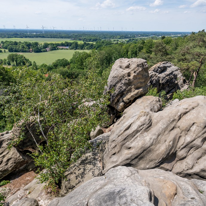 Im Vordergrund große graue Felsen. Dahinter sind grüne Bäume, Wiesen und in der Ferne Wälder und Windräder. (vergrößerte Bildansicht wird geöffnet)