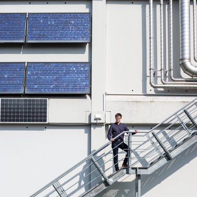 Auf einer Außentreppe steht Wissenschaftler Florian Segger. An der Hauswand hinter ihm sind Rohre und Solarzellen angebracht. (vergrößerte Bildansicht wird geöffnet)