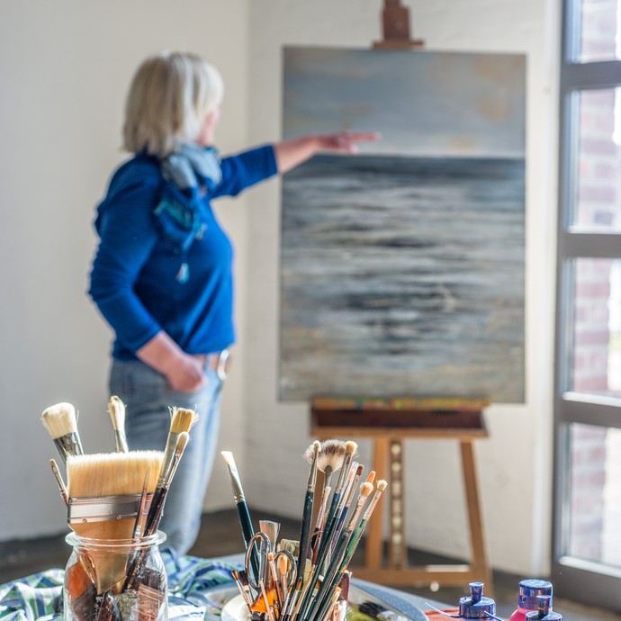 Im Vordergrund sind Pinsel und Farben zum Malen. Im Hintergrund zeigt eine Frau auf ein gemaltes Bild. (vergrößerte Bildansicht wird geöffnet)