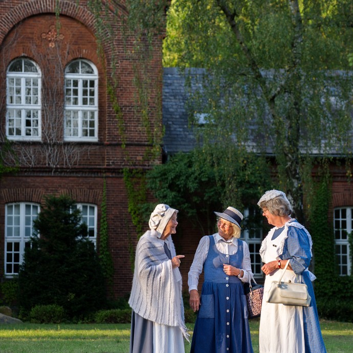 Vor dem alten Klinikgebäude stehen drei Frauen. Sie tragen alte Trachtenkleidung in den Farben blau und weiß. (vergrößerte Bildansicht wird geöffnet)