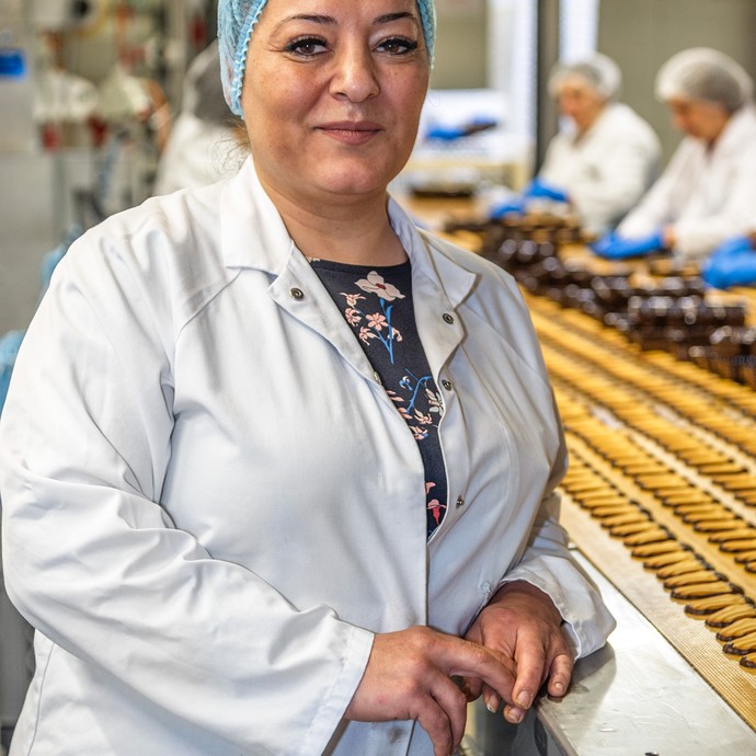 Frau Nasal Ciftci steht am Fließband und lächelt. Sie kümmert sich um die Schokoladenglasur. (vergrößerte Bildansicht wird geöffnet)