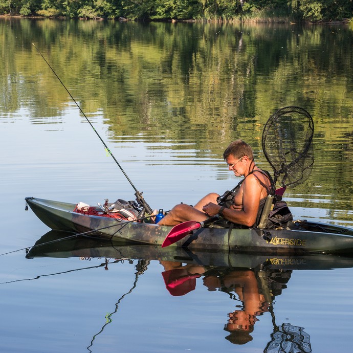 Ein Mann sitzt in einem dunkelgrünen Kanu auf dem See. Er hat eine Angel und einen Kescher dabei. (vergrößerte Bildansicht wird geöffnet)