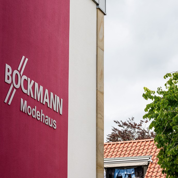 Links ist die Außenfassade mit dem Logo des Modehauses Böckmann. Rechts sind Bäume und das Dach eines Wohnhauses. (vergrößerte Bildansicht wird geöffnet)