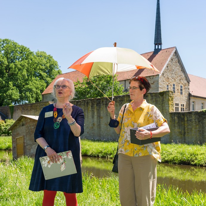 Die Kunstvermittlerinnen Marisa Girard und Annette Hinricher stehen vor dem Kloster. Sie nutzen einen Regenschirm zum Schutz vor der Sonne. (vergrößerte Bildansicht wird geöffnet)