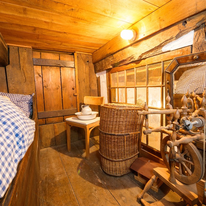 Ein kleiner Raum mit Boden und Wänden aus Holz. Links ist das Bett mit blau-weißer Bettwäsche. (vergrößerte Bildansicht wird geöffnet)