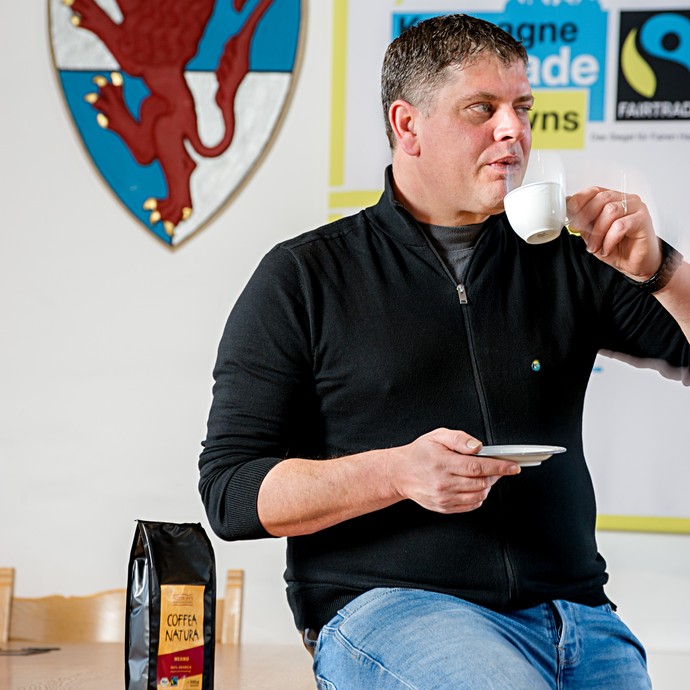 Tobias Meis sitzt auf einem Tisch und trinkt aus einer Tasse Kaffee. Neben ihm steht eine Packung mit "Fairtrade Kaffee". (vergrößerte Bildansicht wird geöffnet)