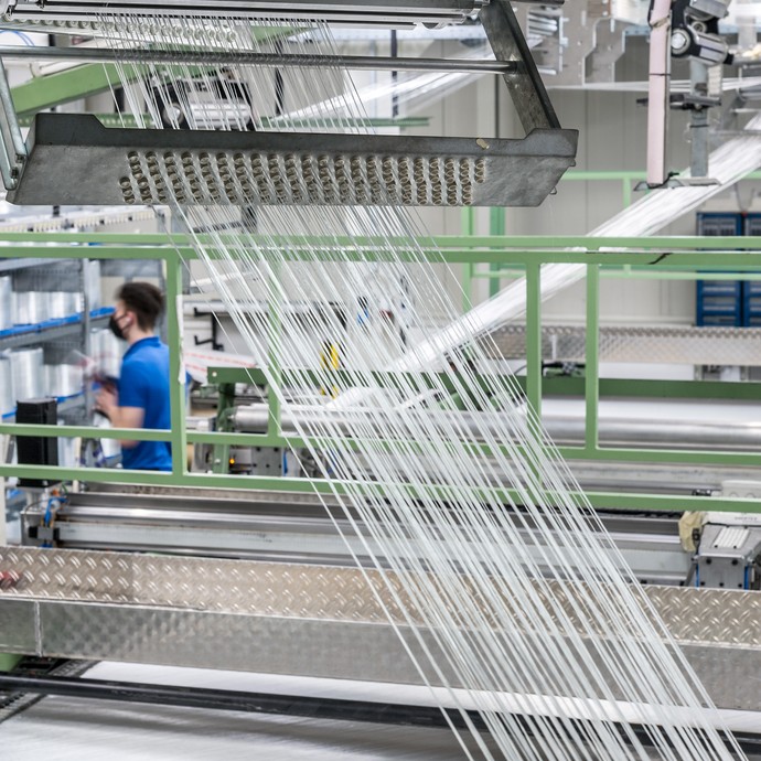 Blick in den Produktionsbereich: Dutzende weiße Garne laufen schräg in eine Maschine gespannt durch das Bild. Im Hintergund steht ein Mitarbeiter. (vergrößerte Bildansicht wird geöffnet)