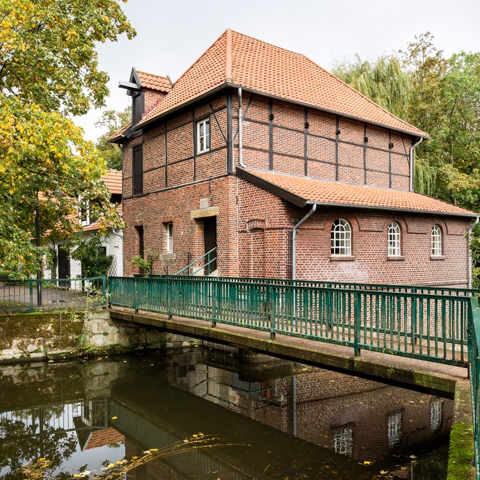 Blick auf eine Brücke. Sie führt über einen Fluss zu einem Gebäude, das aus rötlichen Steinen besteht. (vergrößerte Bildansicht wird geöffnet)