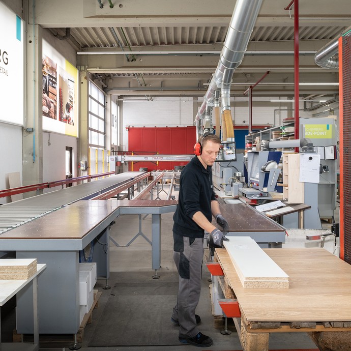 Ein Mitarbeiter steht vor mehreren Tischen mit Holzplatten. Er arbeitet in der Produktionsstraße für Kantenbearbeitung. (vergrößerte Bildansicht wird geöffnet)