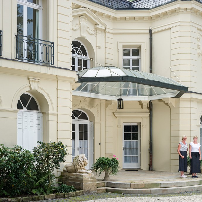 Außenansicht des Landhaus Rothenberge mit cremefarbener Fassade. Vor der Eingangstür stehen drei Frauen mit Schürzen. (vergrößerte Bildansicht wird geöffnet)