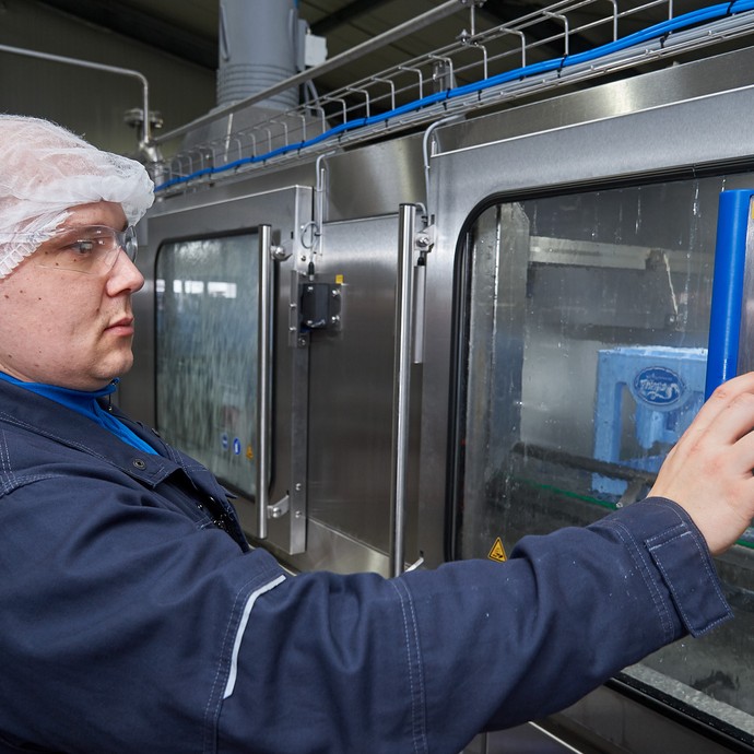 Tim Nokiel bedient ein Kontrollfeld der Waschmaschine. Er trägt ein Haarnetz für die Hygiene am Arbeitsplatz. (vergrößerte Bildansicht wird geöffnet)