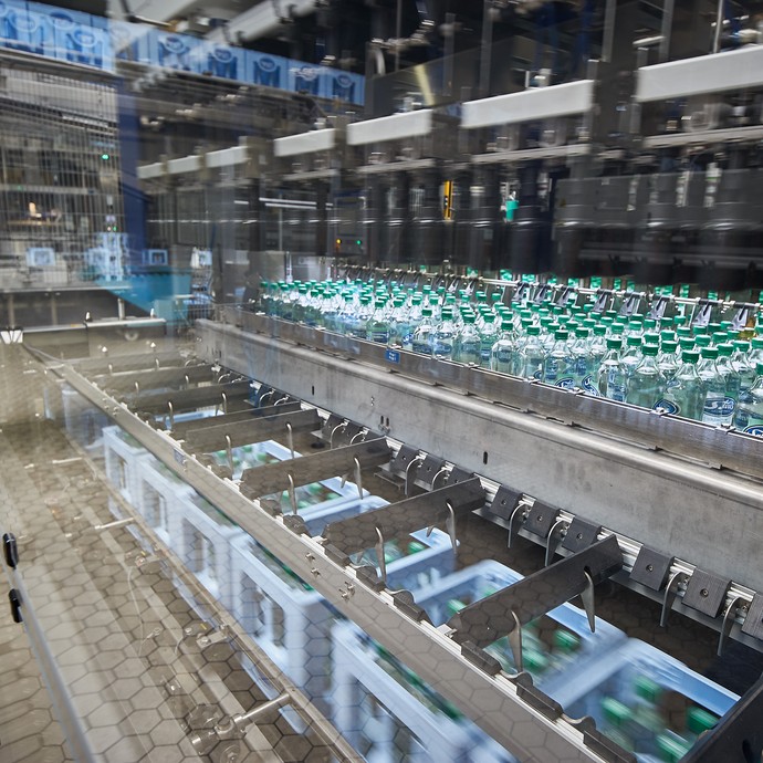 Im Vordergrund ist eine große Maschine. Sie füllt die Wasserflaschen in hellblaue Kisten. Links im Hintergrund steht ein Arbeiter. (vergrößerte Bildansicht wird geöffnet)