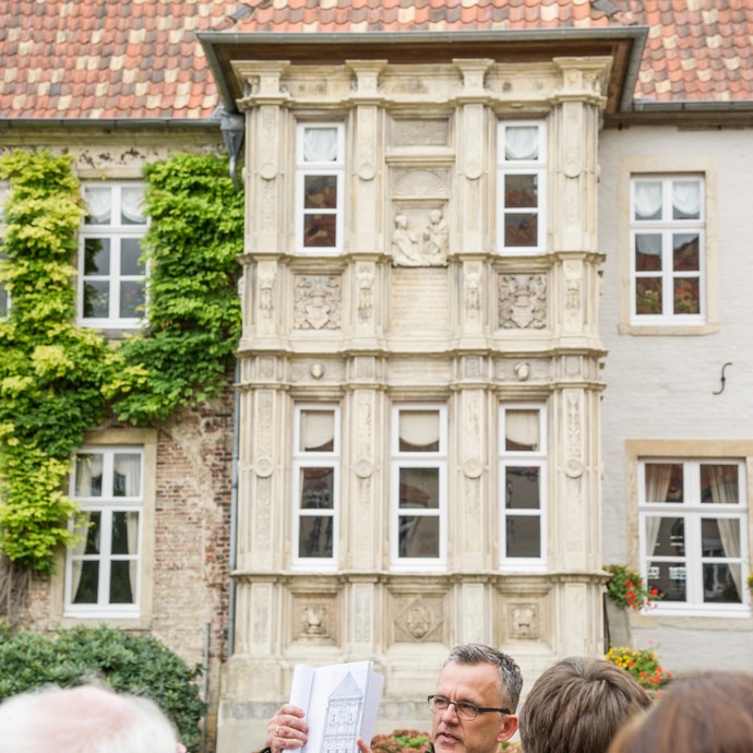 Ein Mann hält eine Zeichnung von einem Gebäude hoch. Vor ihm stehen Personen, die ihm zuhören. Dahinter ist das Gebäude. (vergrößerte Bildansicht wird geöffnet)