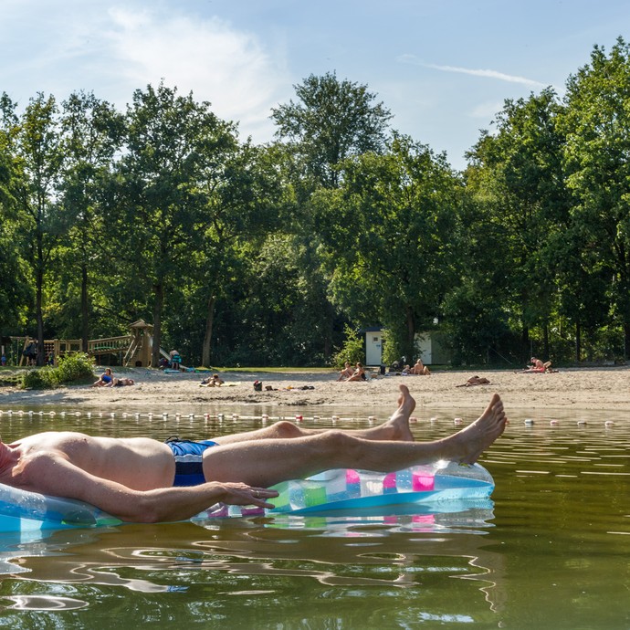 Im Vordergrund liegt ein Mann in Badehose auf einer hellblauen Luftmatratze auf dem See. Am Strand spielen Kinder und Erwachsene sonnen sich. (vergrößerte Bildansicht wird geöffnet)